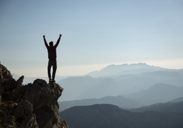 person standing on edge of cliff with arms raised to the sky